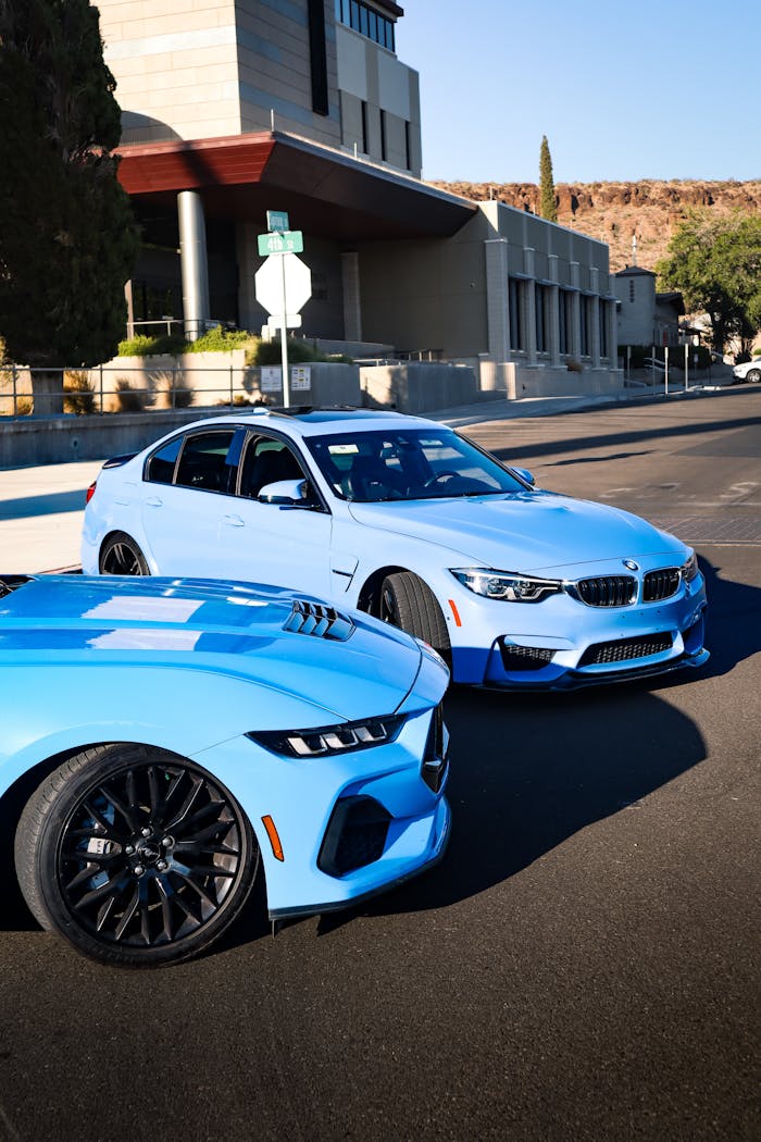 Contact Two blue sports cars parked on a sunny urban street in front of modern architecture.
