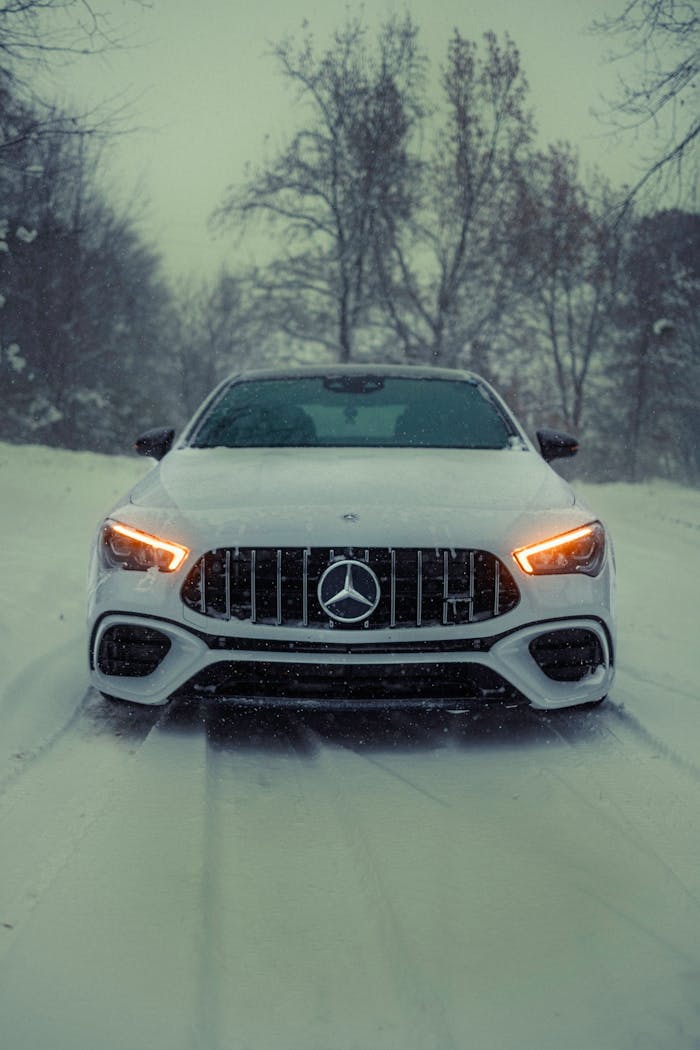 Front view of a luxury car on a snowy road, headlights glowing in winter setting.